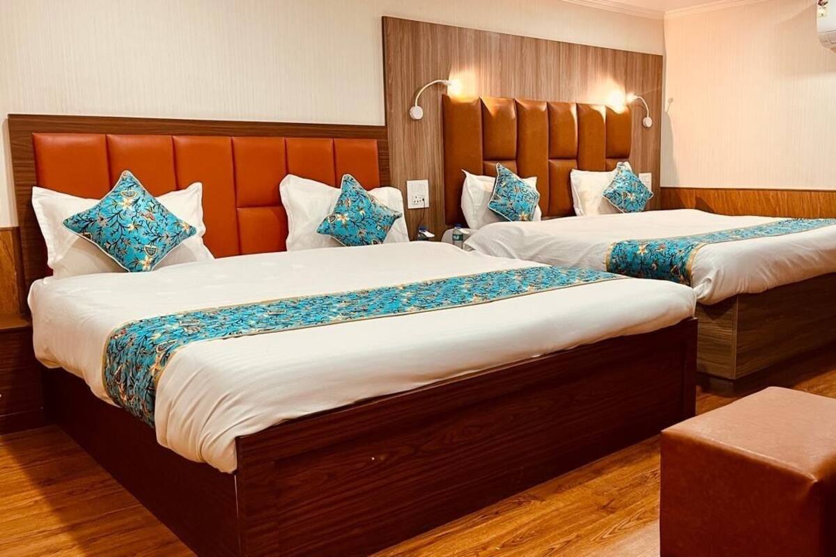 A hotel room with two beds, each with a wooden headboard, white bedding, and blue patterned throw blankets. There is a wooden bench and a wall-mounted air conditioning unit.