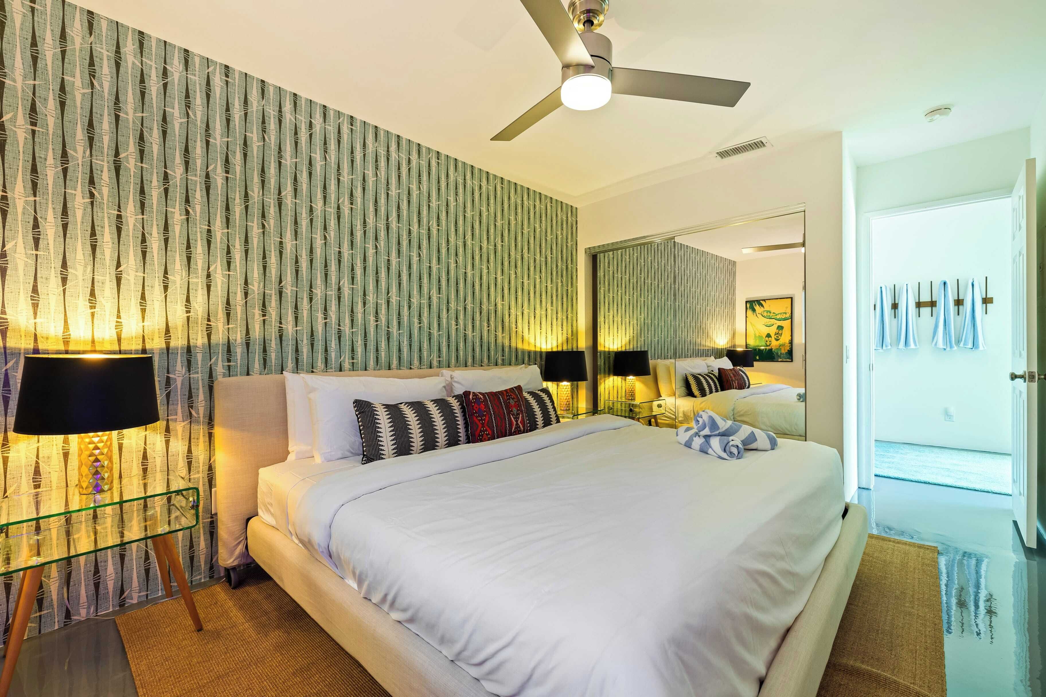A modern hotel room with a large bed, a ceiling fan, a nightstand with lamps, and a large mirror reflecting the room.
