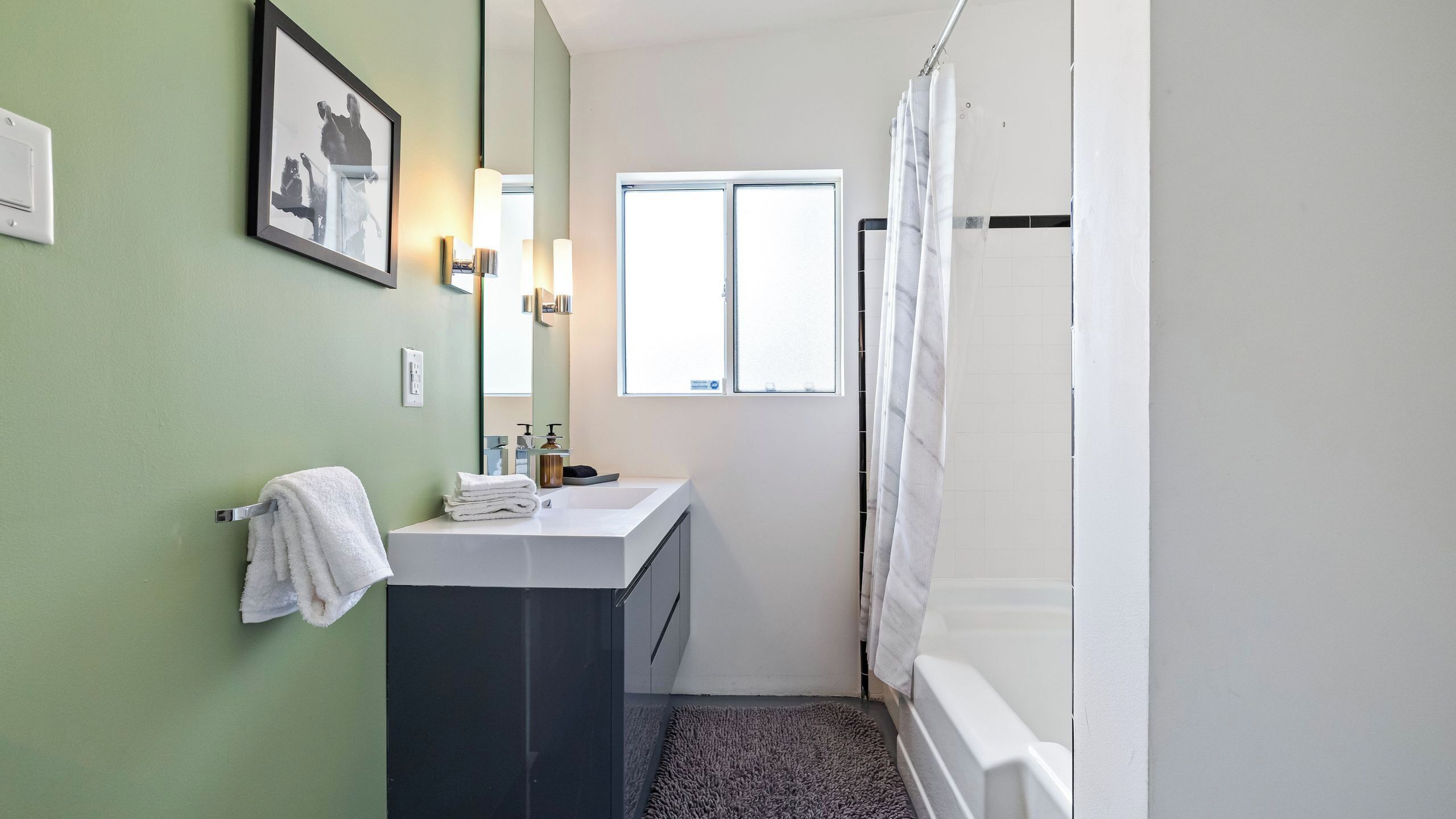 A bathroom with a white sink, dark cabinet, green walls, a framed picture, and a window.