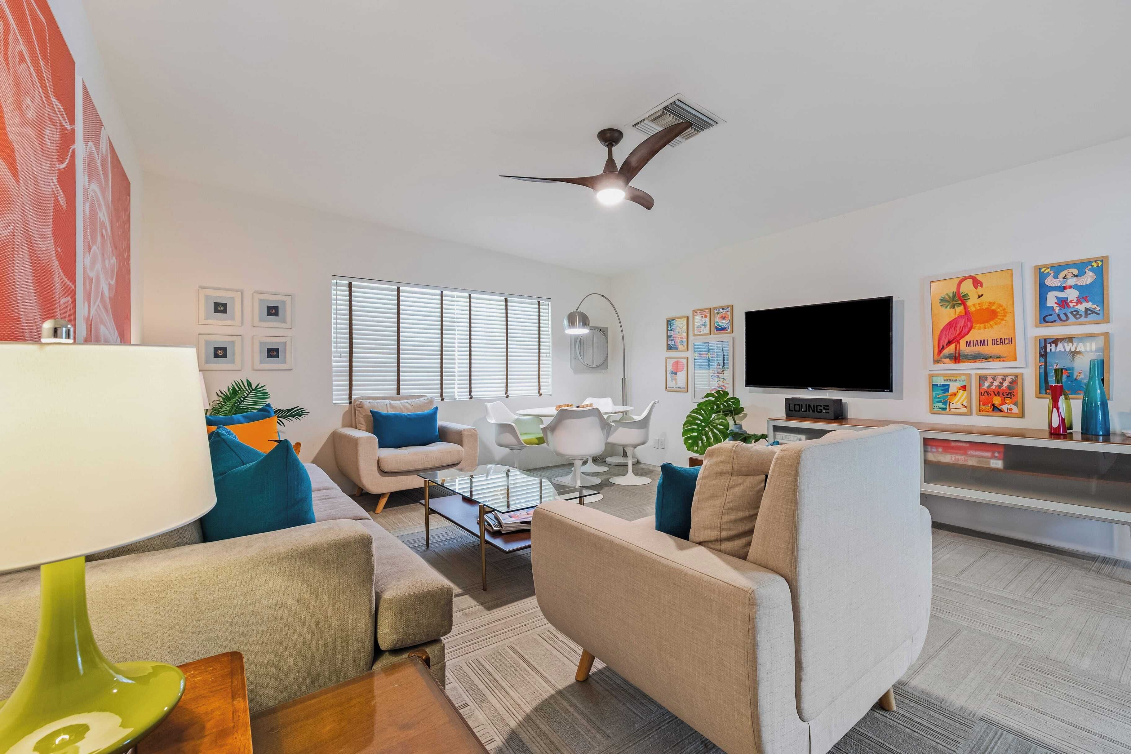A modern living room with a sofa, armchairs, a coffee table, and a TV. There is a ceiling fan, a window with blinds, and a wall with framed pictures.