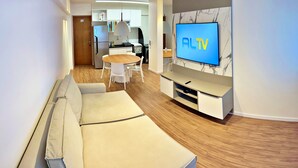 Moderni olohuone, jossa on sohva, seinälle asennettu televisio, ruokailutila pöytineen ja tuoleineen sekä taustalla keittiö.