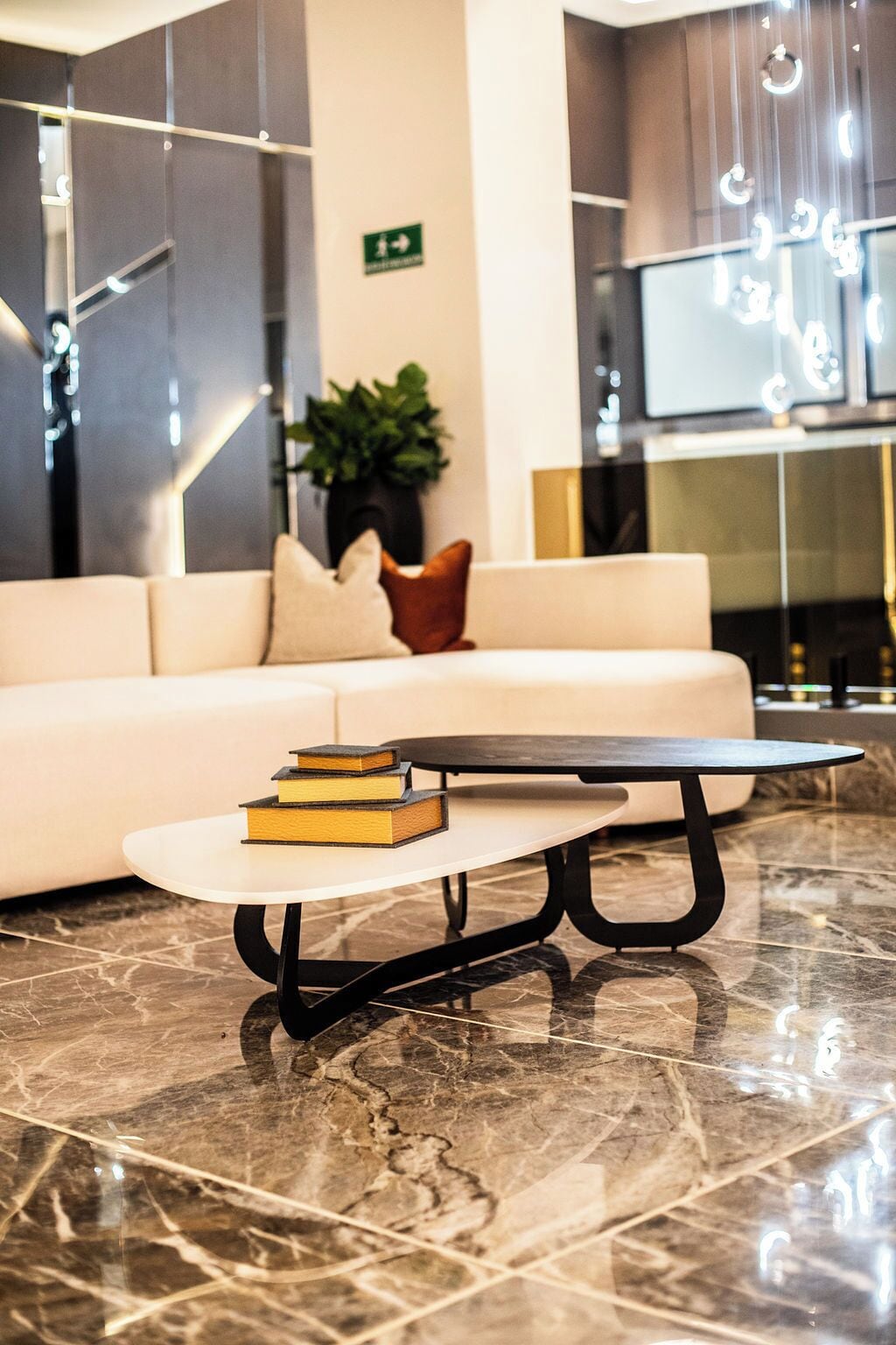 Un vestíbulo moderno con piso de mármol, un sofá blanco con cojines, una pequeña mesa redonda con libros y una señal de salida verde en la pared.