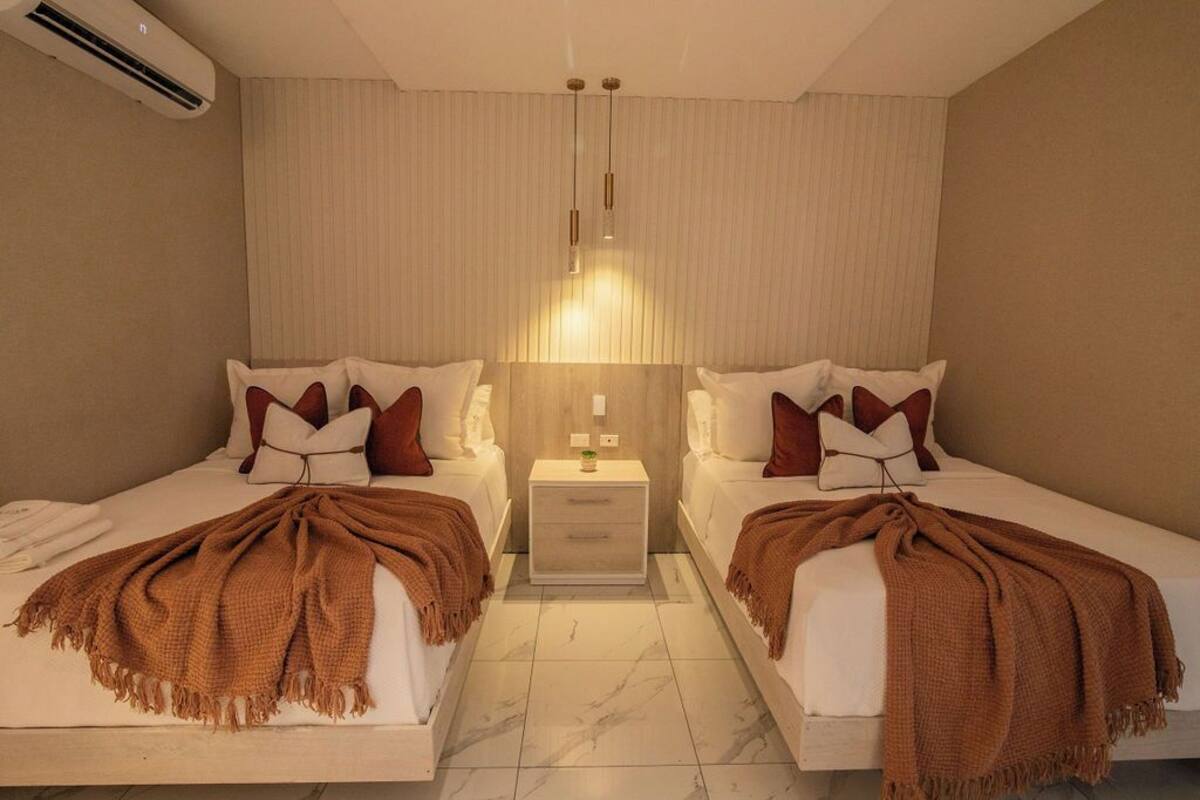 Habitación de hotel con dos camas, una mesita de noche, una lámpara y aire acondicionado instalado en la pared.