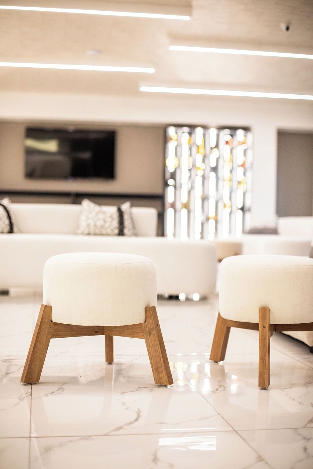 Una sala de estar moderna con un sofá blanco, dos otomanas blancas con patas de madera y un televisor montado en la pared.