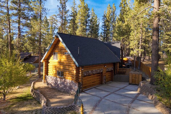 A log cabin with a stone foundation, a garage, and a patio area surrounded by trees.