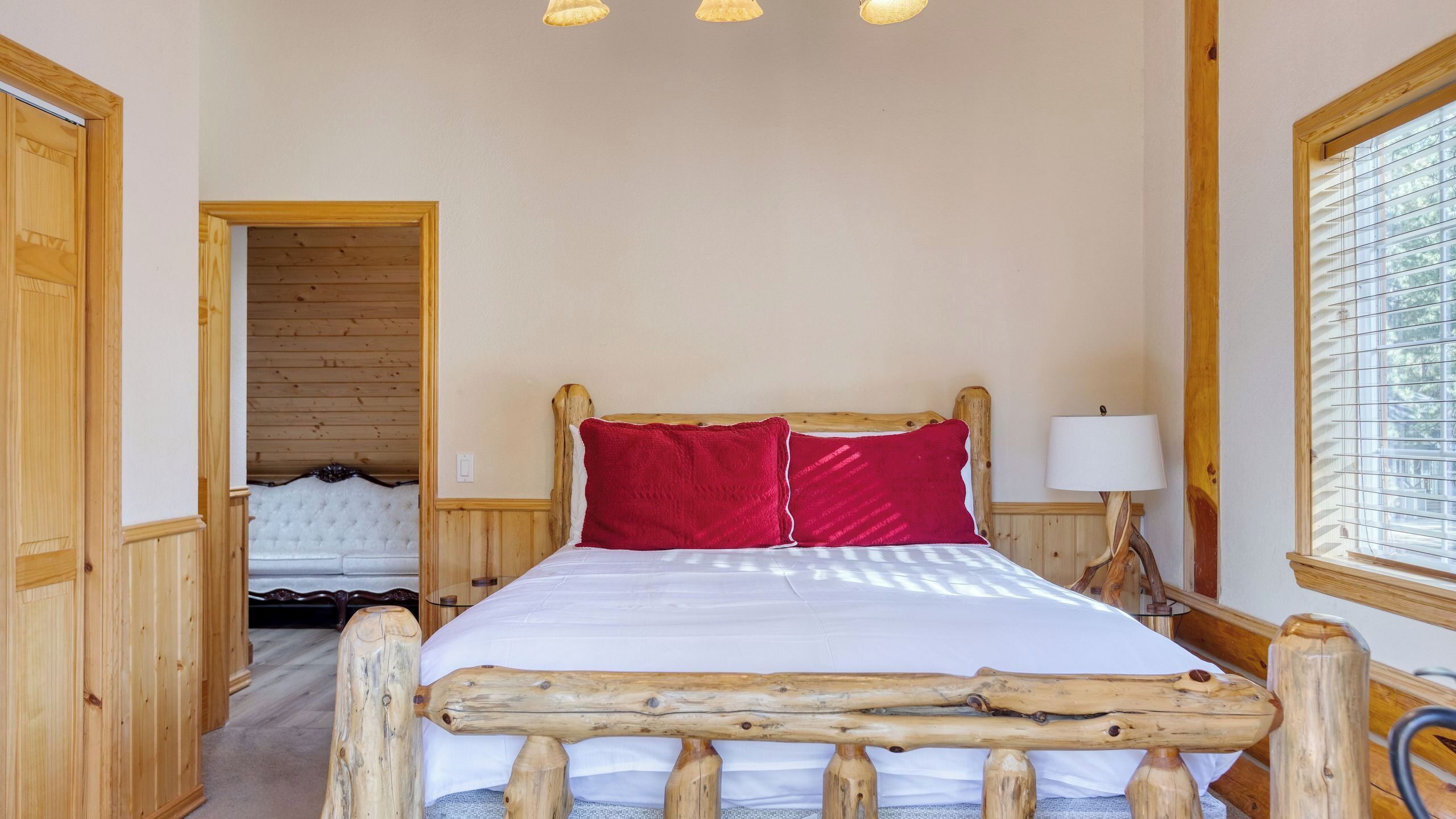 A wooden-framed bed with a white mattress and red pillows, a bedside lamp, and a window with blinds.