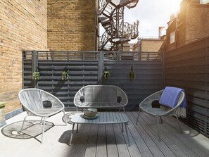 Une terrasse avec deux chaises en osier blanc, une petite table ronde et une clôture en métal gris ornée de plantes suspendues.