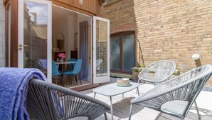 Un patio al aire libre con sillas de mimbre y una mesa, una puerta corrediza de vidrio que da a un espacio interior y un muro de ladrillo.
