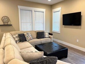 A living room with a sectional sofa, a black ottoman, and a flat-screen TV mounted on the wall.