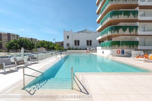 Ein Swimmingpool mit klarem, blauem Wasser, umgeben von Liegestühlen und einem weißen Gebäude mit Balkonen im Hintergrund.