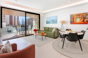 A modern living room with a green sofa, a dining table with chairs, a coffee table with fruit, and a painting on the wall.