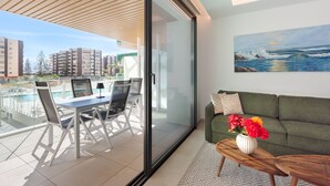 Ett modernt vardagsrum med en grön soffa, ett soffbord med en vas med blommor, en matplats med bord och stolar samt en stor glasdörr som leder ut till en balkong med pool och utsikt över staden.