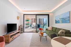 Ein modernes Wohnzimmer mit einem Flachbildfernseher, einem grünen Sofa, einem Couchtisch mit Blumenvase und einem großen Fenster mit Blick auf einen Pool und Gebäude.