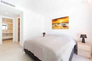 Ein Schlafzimmer mit einem Bett, Nachttischen, einem Bild an der Wand und einem durch eine offene Tür einsehbaren Badezimmer.