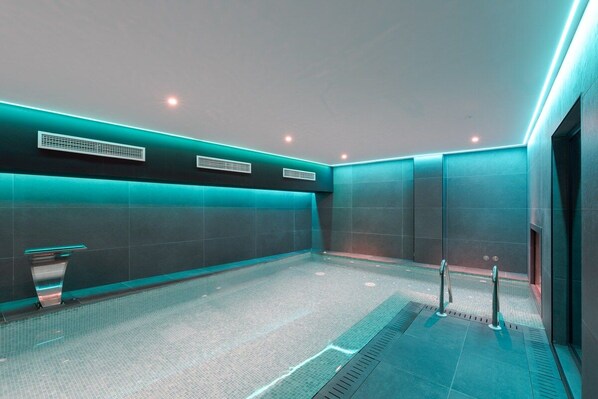 Un área de piscina interior moderna con suelo de baldosas, iluminación empotrada y una pared de acento color turquesa.