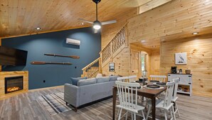 A cozy cabin interior with a fireplace, a sofa, a dining table with chairs, and a staircase.
