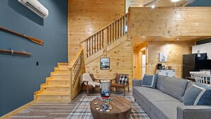 A cozy cabin interior with a wooden staircase, a living area with a sofa and chairs, a dining table, and a kitchen area.