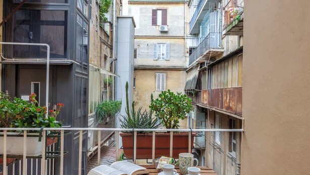A narrow urban courtyard with a wooden table, two cups, a book, and potted plants.