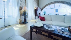 A cozy living room with a white sofa, a coffee table, and a lamp. There is a stone wall and a large window with curtains.