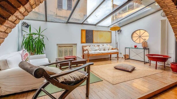 A spacious living area with a high ceiling, a skylight, a sofa, a coffee table, a red bathtub, and a large plant.