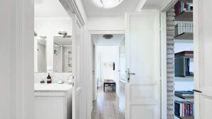 A hallway with a bathroom on the left, a door leading to a room on the right, and a bookshelf in the background.