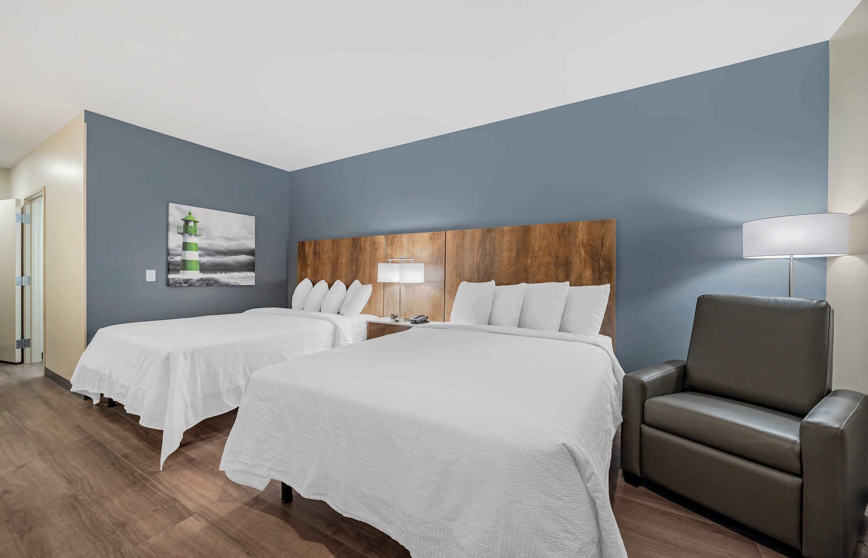 A hotel room with two beds, a wooden headboard, a chair, a lamp, and a painting of a lighthouse.