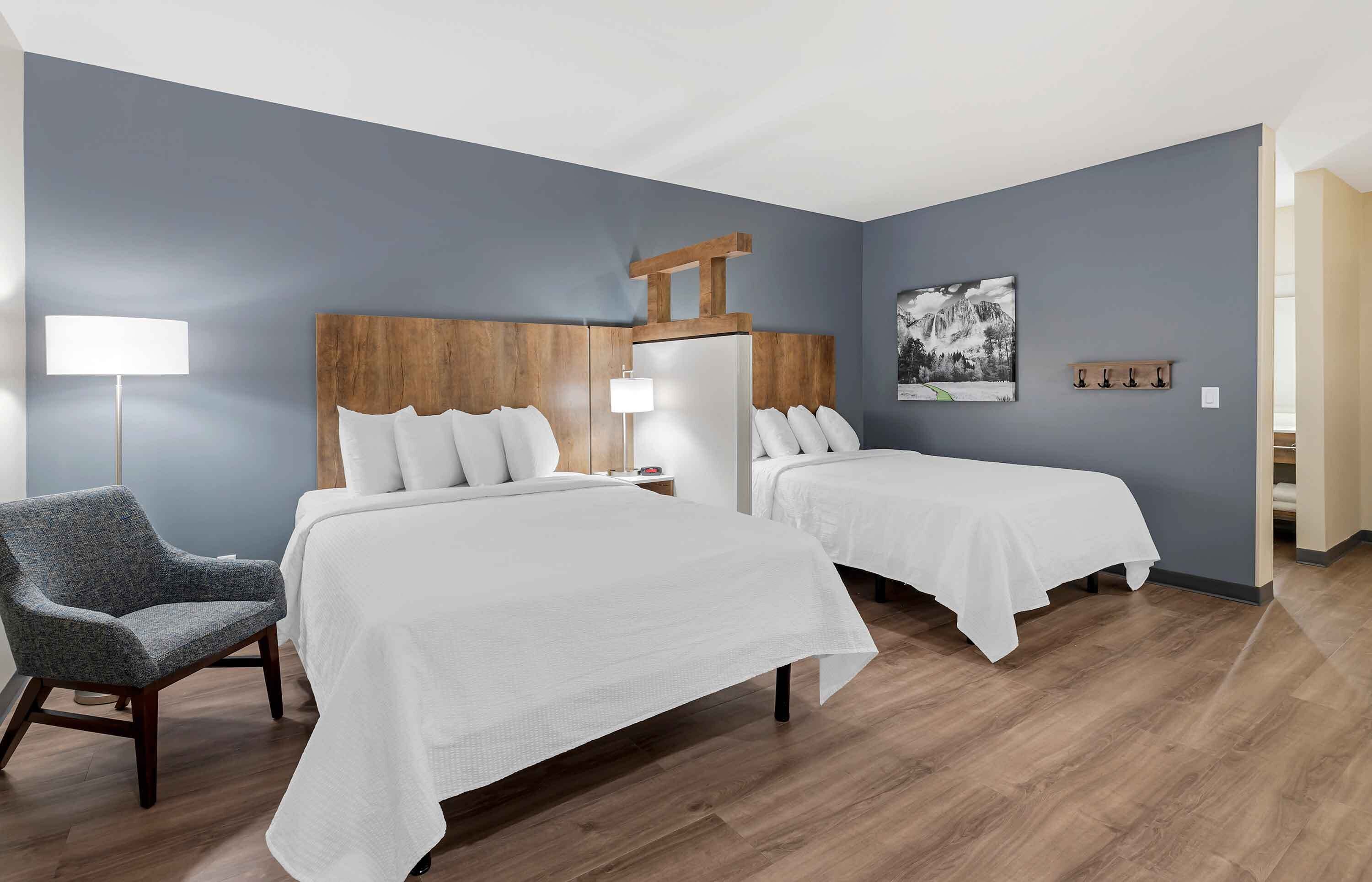 Habitación de hotel con dos camas, una silla, una mesita de noche, una lámpara y un perchero en la pared.