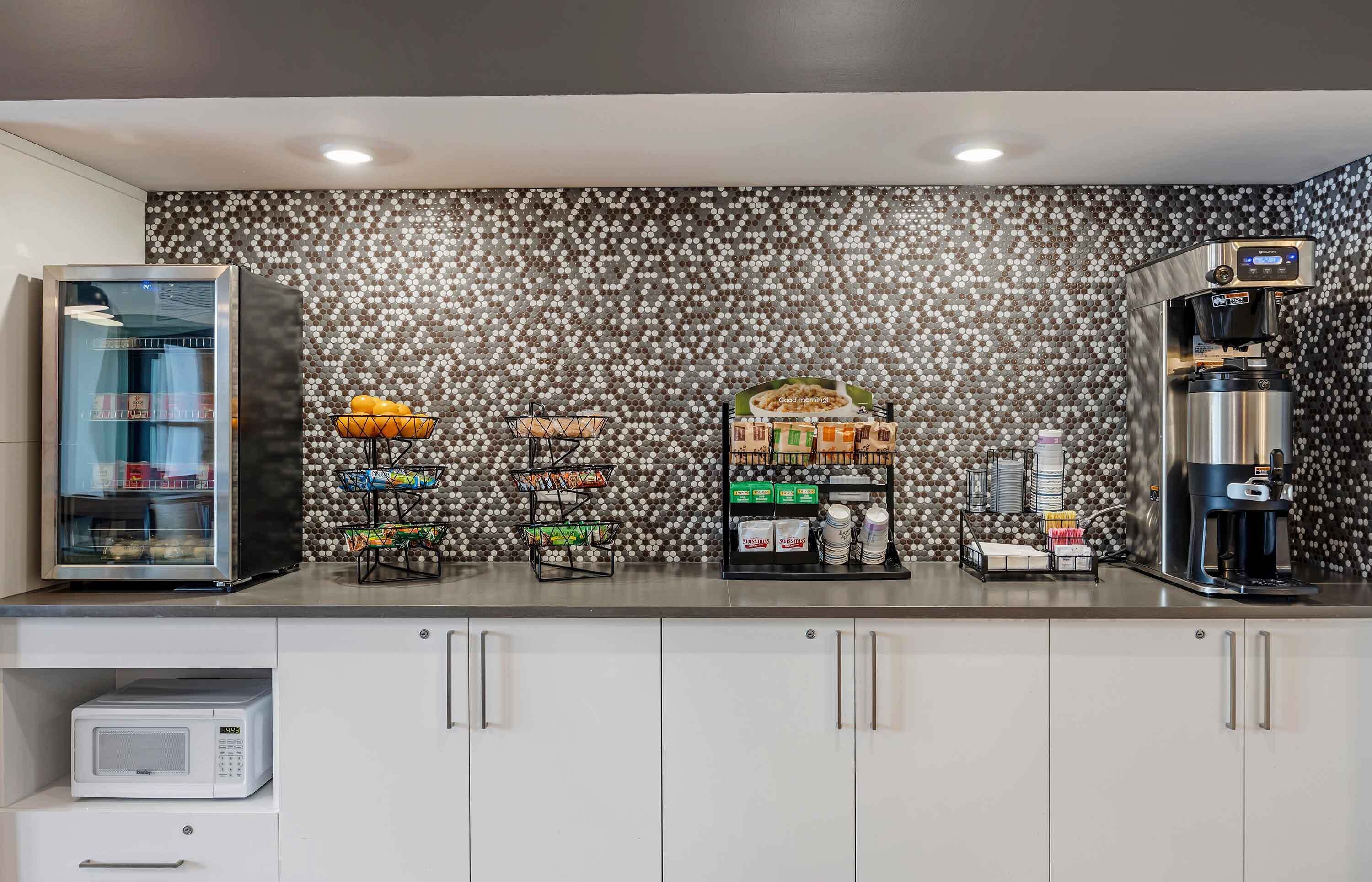 A kitchenette with a microwave, a coffee machine, and a refrigerator. There are baskets with fruits and a shelf with packaged food items.