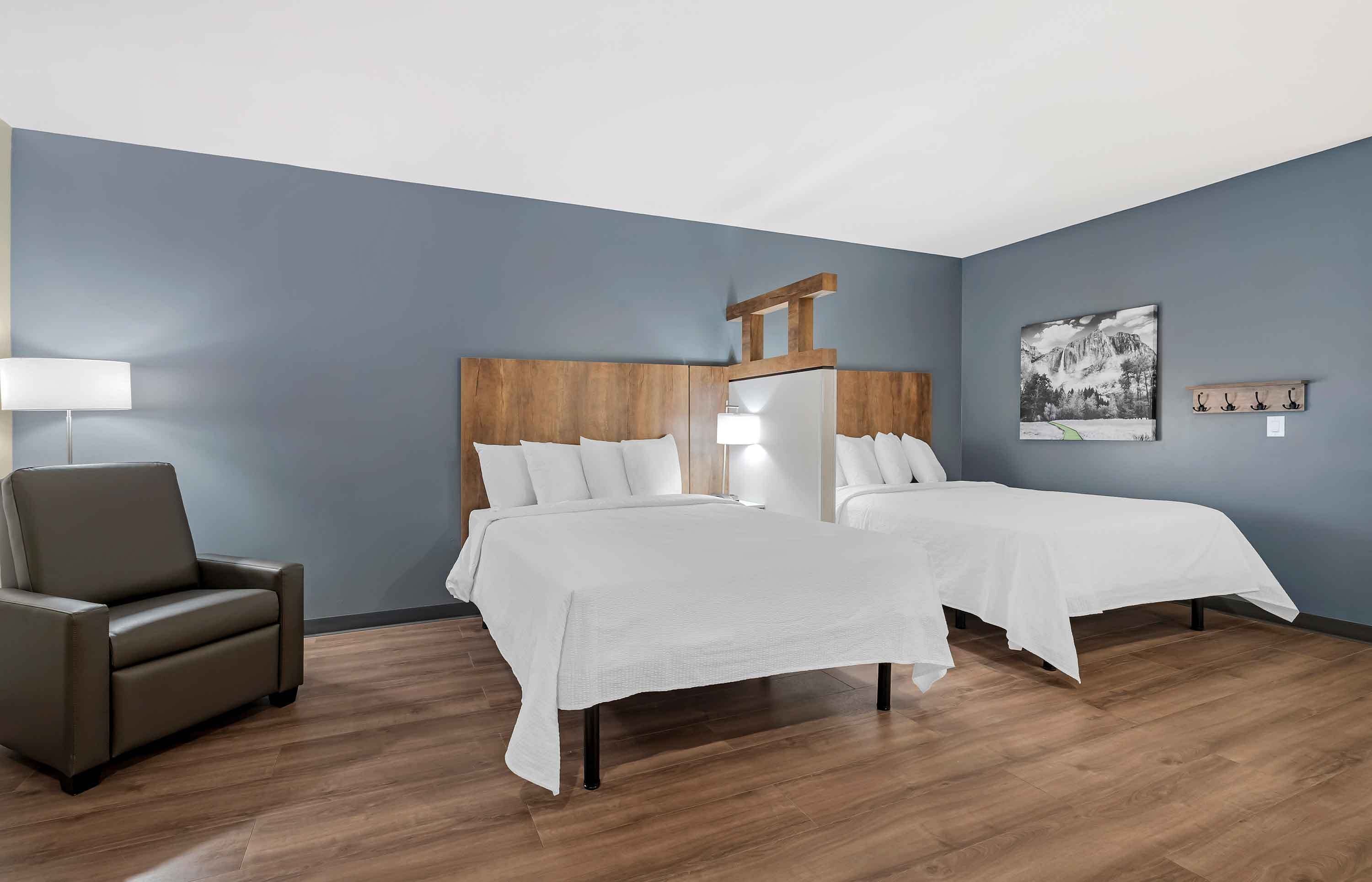 A hotel room with two beds, a wooden headboard, a chair, a lamp, and a painting on the wall.