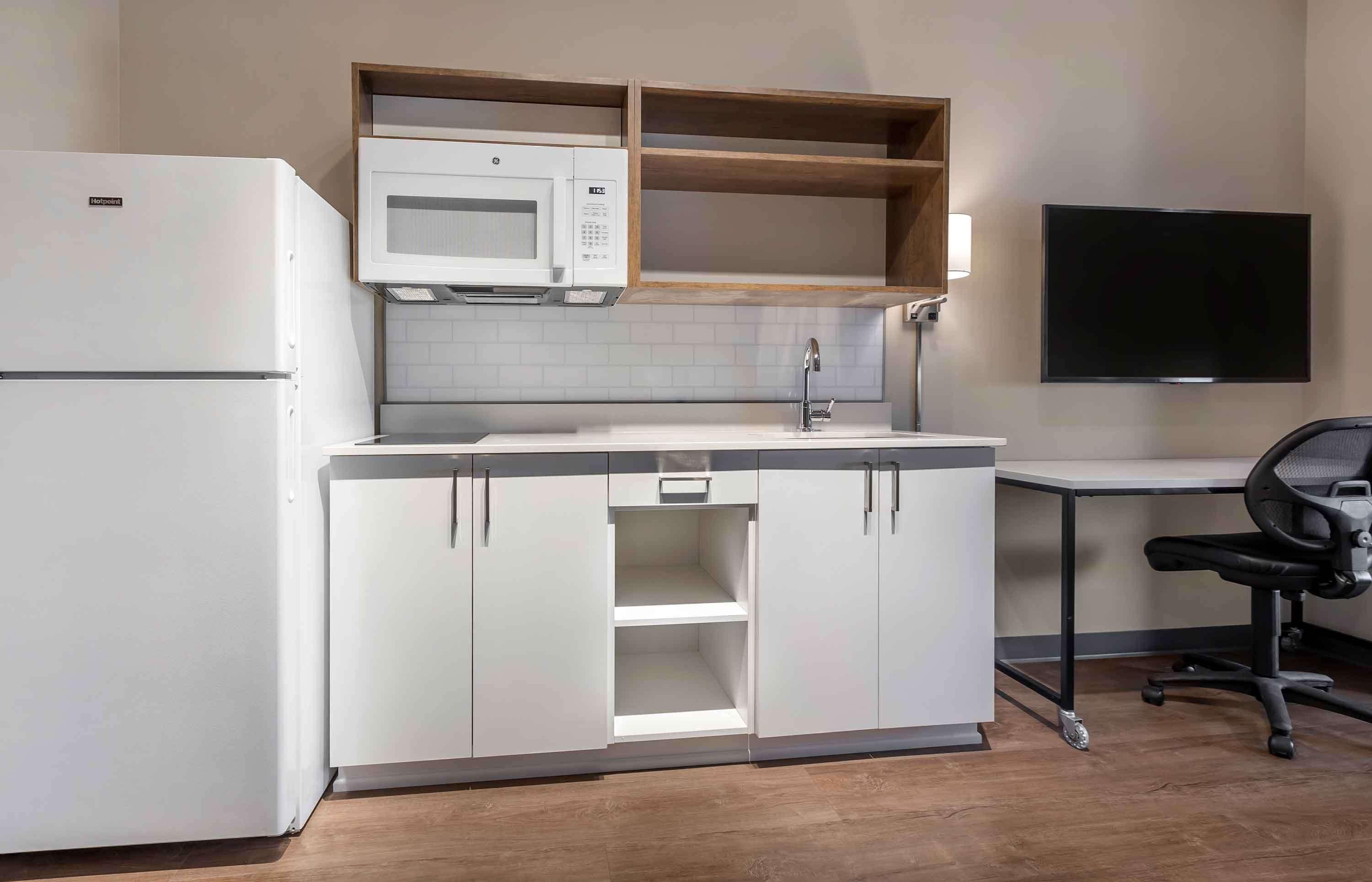 A compact kitchen with a microwave, a refrigerator, and a sink. There is a wooden shelf above the microwave and a flat-screen TV mounted on the wall.