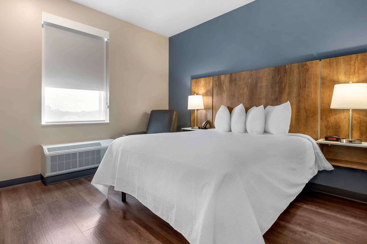 A hotel room with a large bed, a wooden headboard, two bedside tables with lamps, a window with blinds, and a wall-mounted heater.