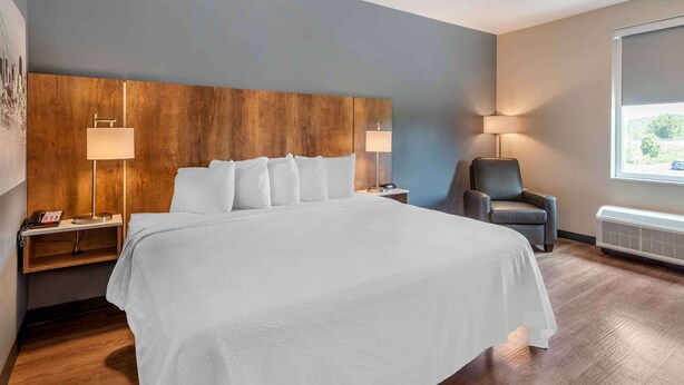 A modern hotel room with a large bed, wooden headboard, two bedside tables with lamps, a chair, and a window with a view. - Extended Stay America Premier Suites - San Antonio - Schertz (Schertz)