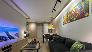 Ein modernes Wohnzimmer mit einem großen Flachbildfernseher, einem schwarzen Sofa mit grünen Kissen, einem Holztisch und einer Küche im Hintergrund.