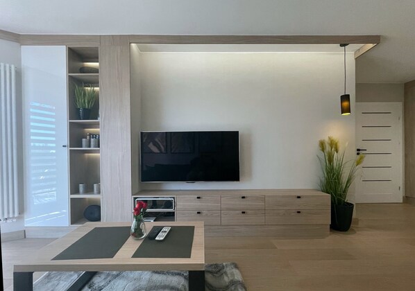 A modern living room with a flat-screen TV mounted on the wall, a wooden coffee table with a vase of flowers, and a potted plant.