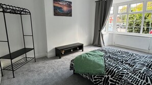 A bedroom with a bed, a black metal shelf, a small cabinet, a painting, and a window with curtains.