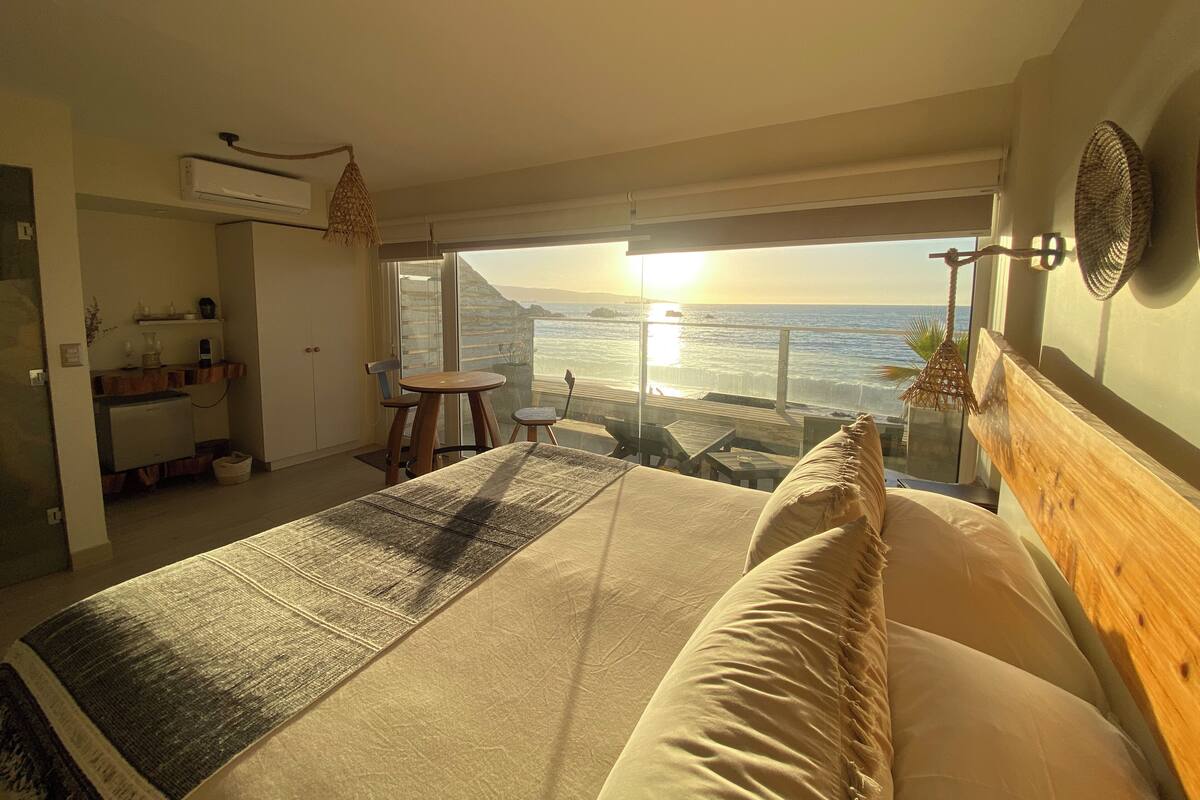 Un dormitorio con una cama grande, una mesita, una silla, un balcón con vista al mar y una pequeña cocina.
