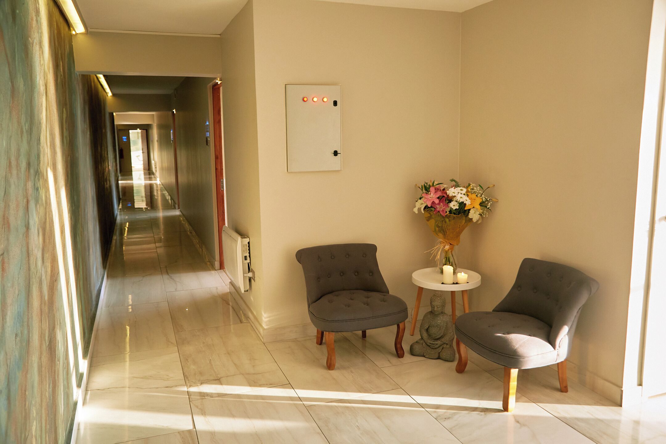 A hotel lobby with a marble floor, two grey armchairs, a small round table with a vase of flowers, and a wall-mounted light switch.