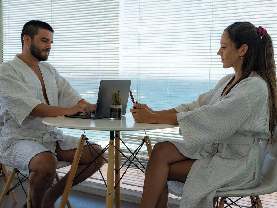 Two people in white robes sitting at a round table with a laptop, a small plant, and a glass of water. They are in a room with large windows and blinds.
