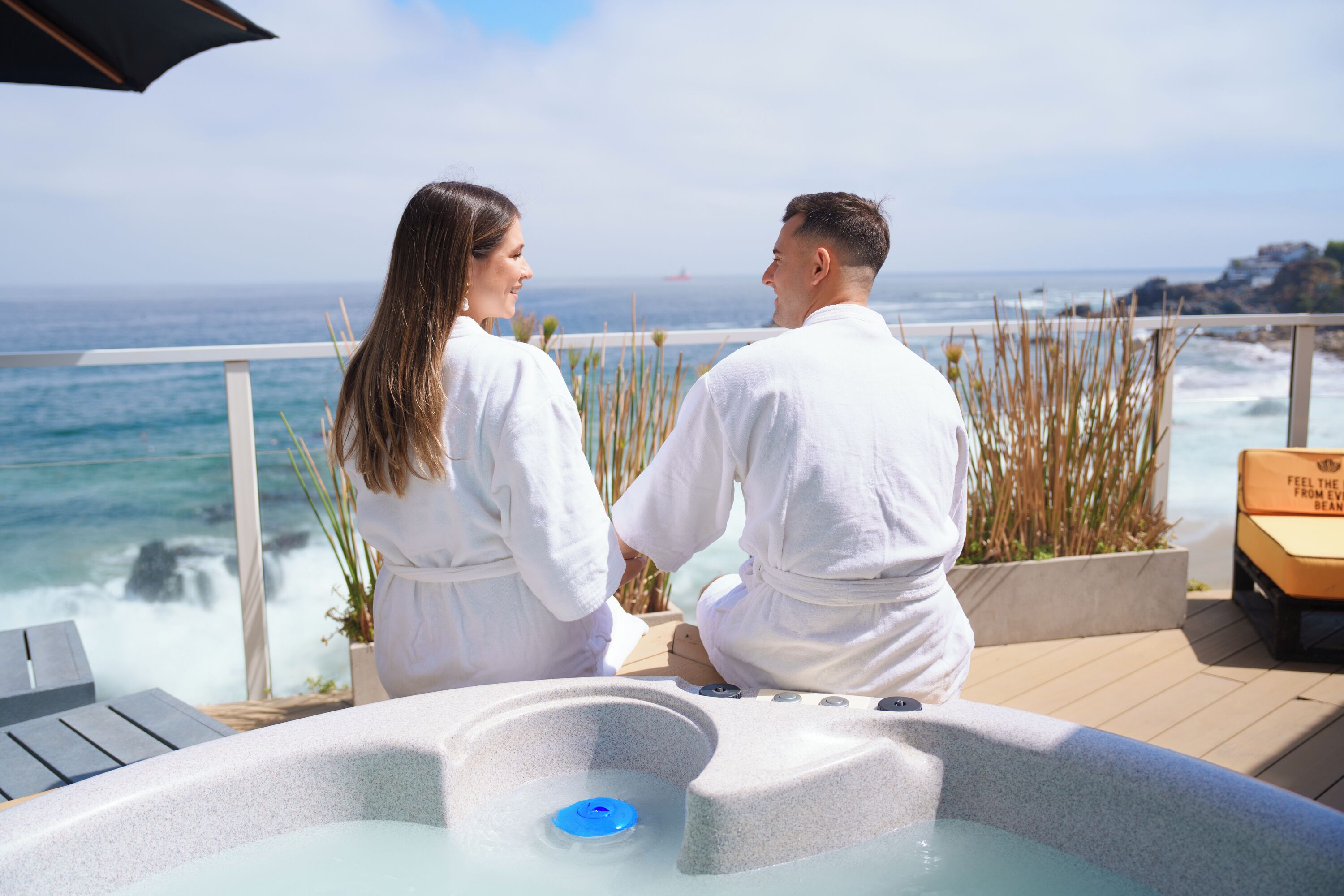 A couple in white robes by a hot tub overlooking the ocean.