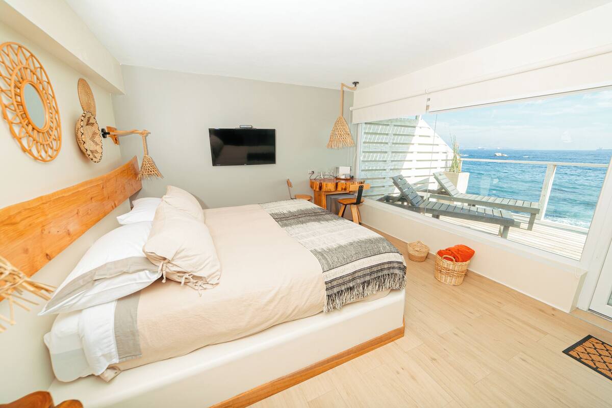 Un dormitorio con una cama grande, televisión, cabecero de madera, vista al océano y un balcón con una silla.