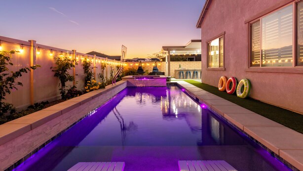 A swimming pool with a deck, surrounded by a fence with string lights, and a building with a window and a door.