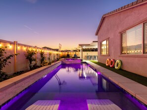 A swimming pool with a deck, surrounded by a fence with string lights, and a building with a window and a door.