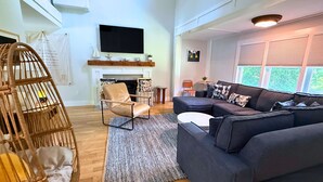 Un salon comprenant un canapé, un fauteuil, une cheminée, un téléviseur et une fenêtre avec des stores.