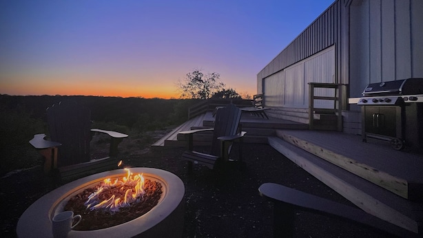 Un patio exterior con un brasero, asientos y una parrilla, con vistas a un paisaje montañoso al atardecer.