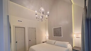 Un dormitorio con una cama grande, un candelabro, dos puertas, una mesita de noche y una lámpara de pared.