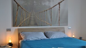 A bedroom with a bed, a nightstand, a wall-mounted lamp, and a large painting of a bridge.