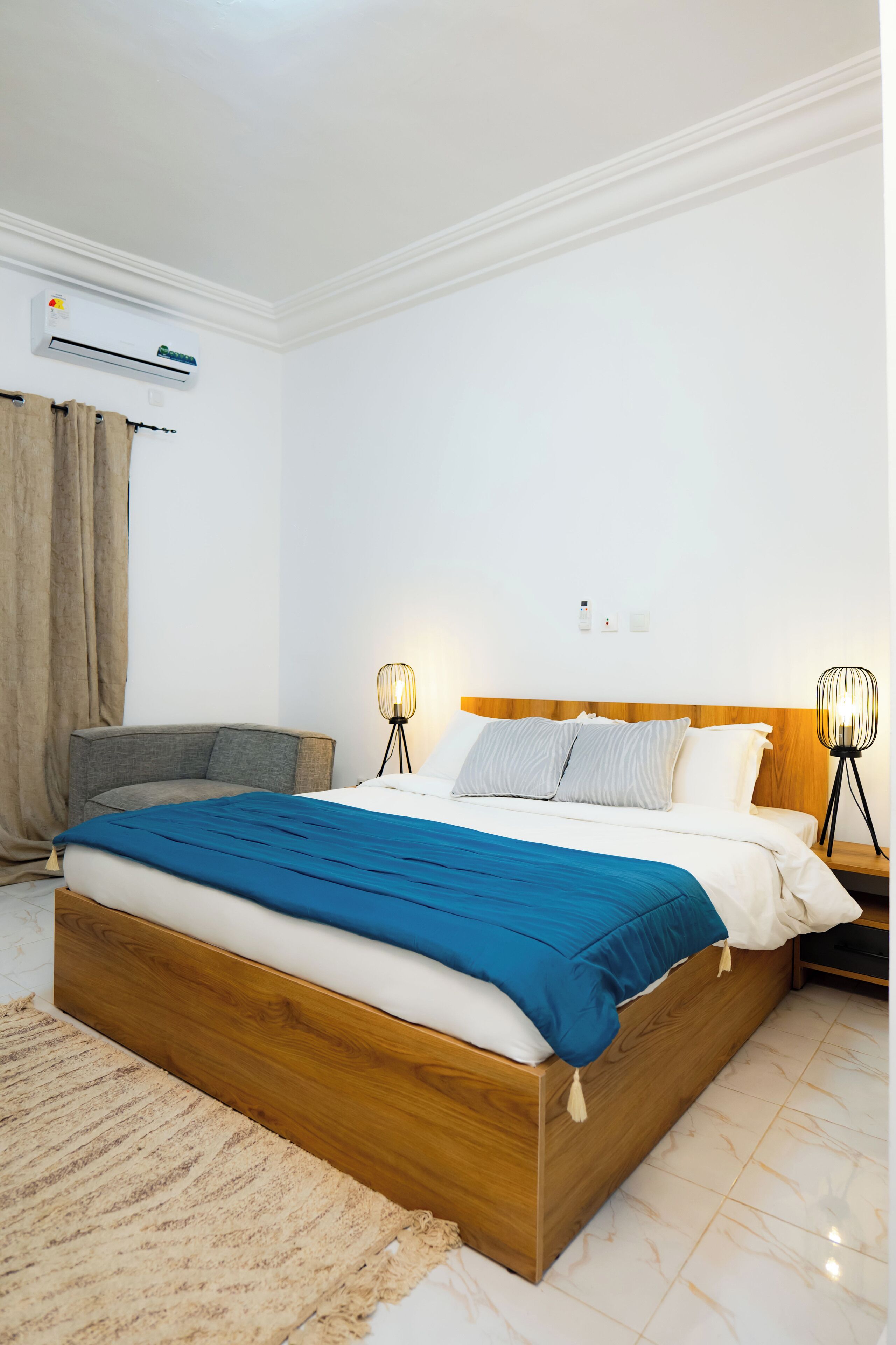 A modern hotel room with a wooden bed, a grey armchair, a beige rug, and two bedside lamps.