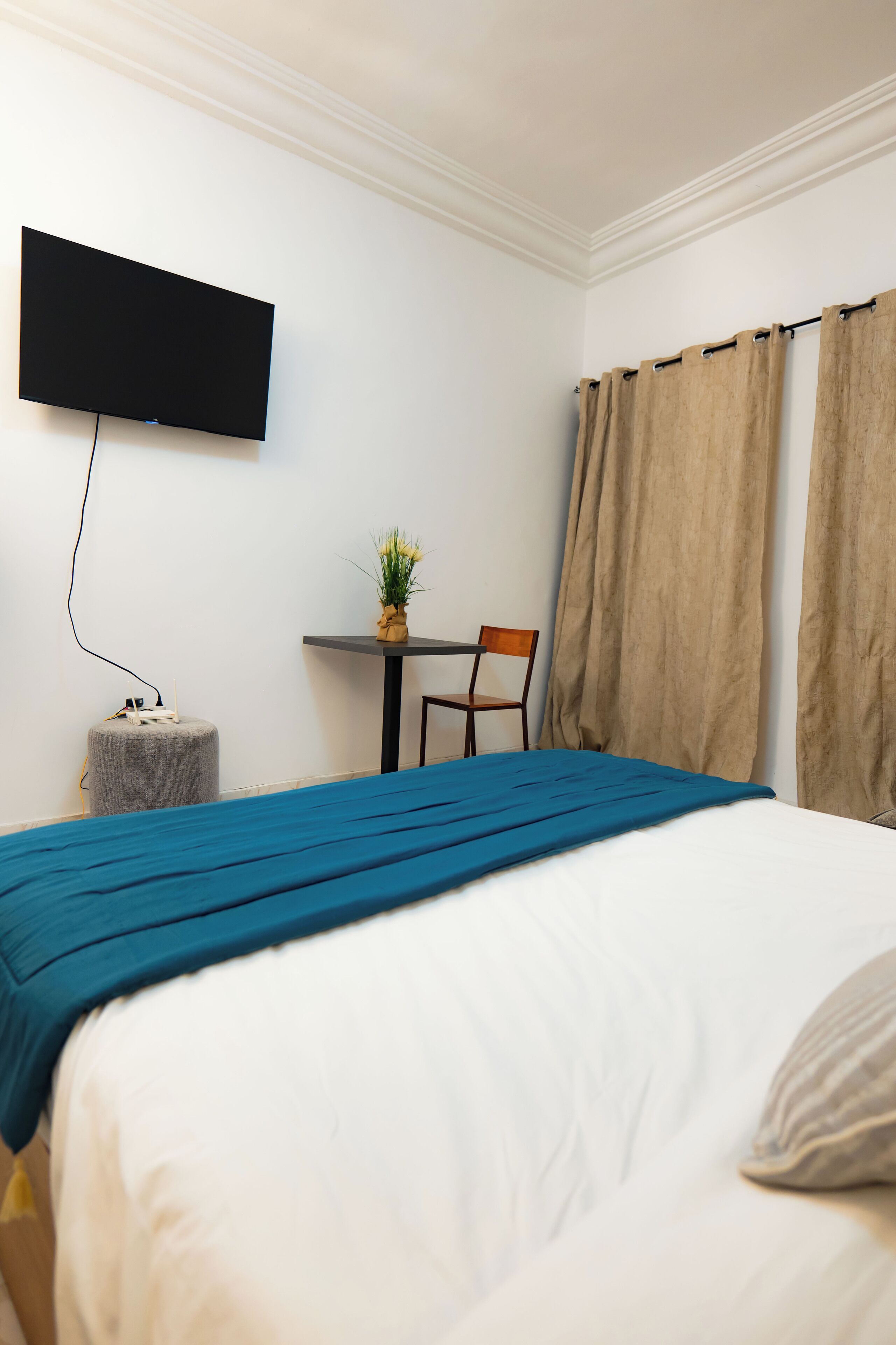 Une chambre d’hôtel avec un lit, un téléviseur à écran plat fixé au mur, une petite table avec une chaise et une fenêtre avec des rideaux.