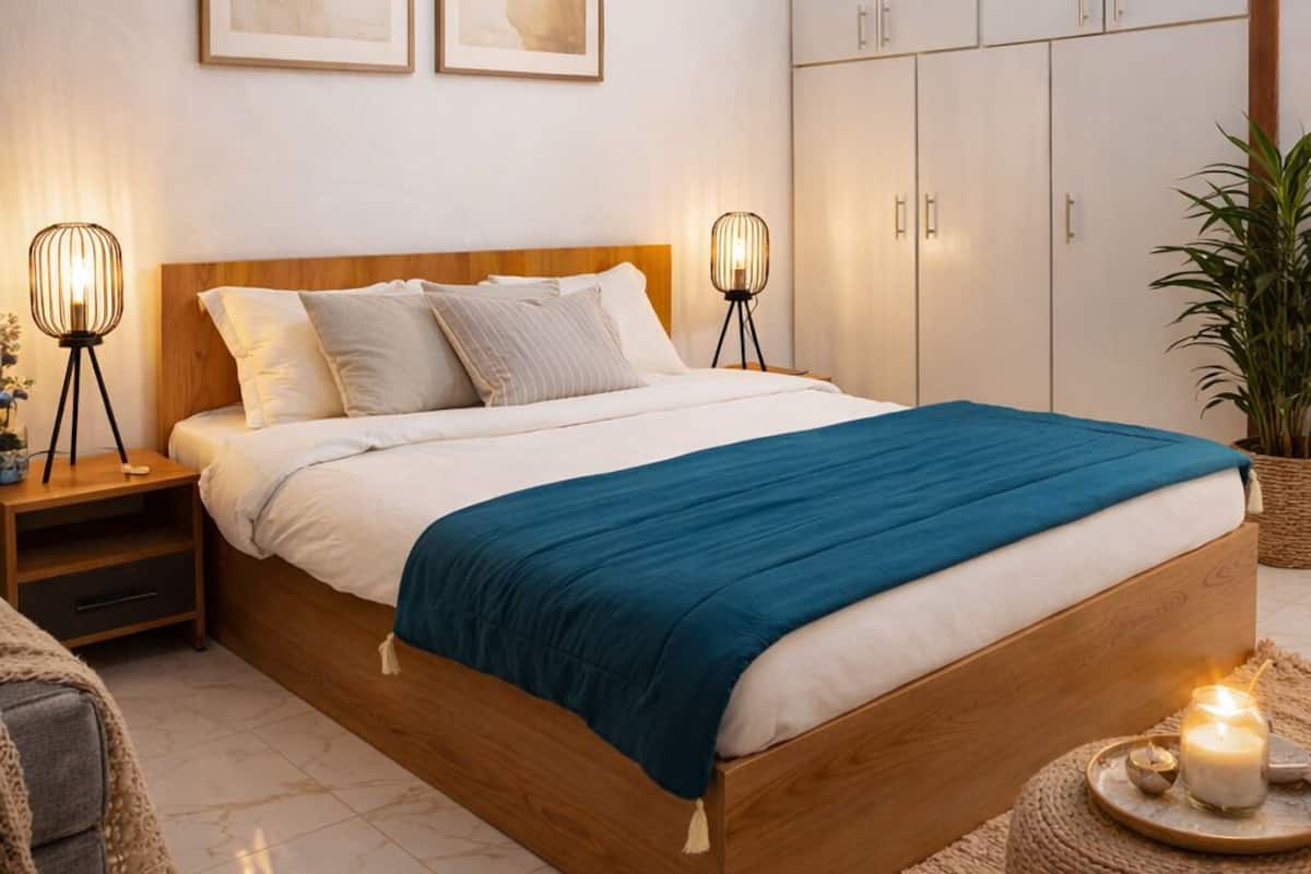 A bedroom with a wooden bed, two bedside lamps, a blue bedspread, a large wardrobe, and two abstract paintings on the wall.
