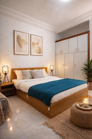 A bedroom with a wooden bed, two bedside lamps, a blue bedspread, a large wardrobe, and two abstract paintings on the wall. - Casa Hôtel Résidence (Abidjan)